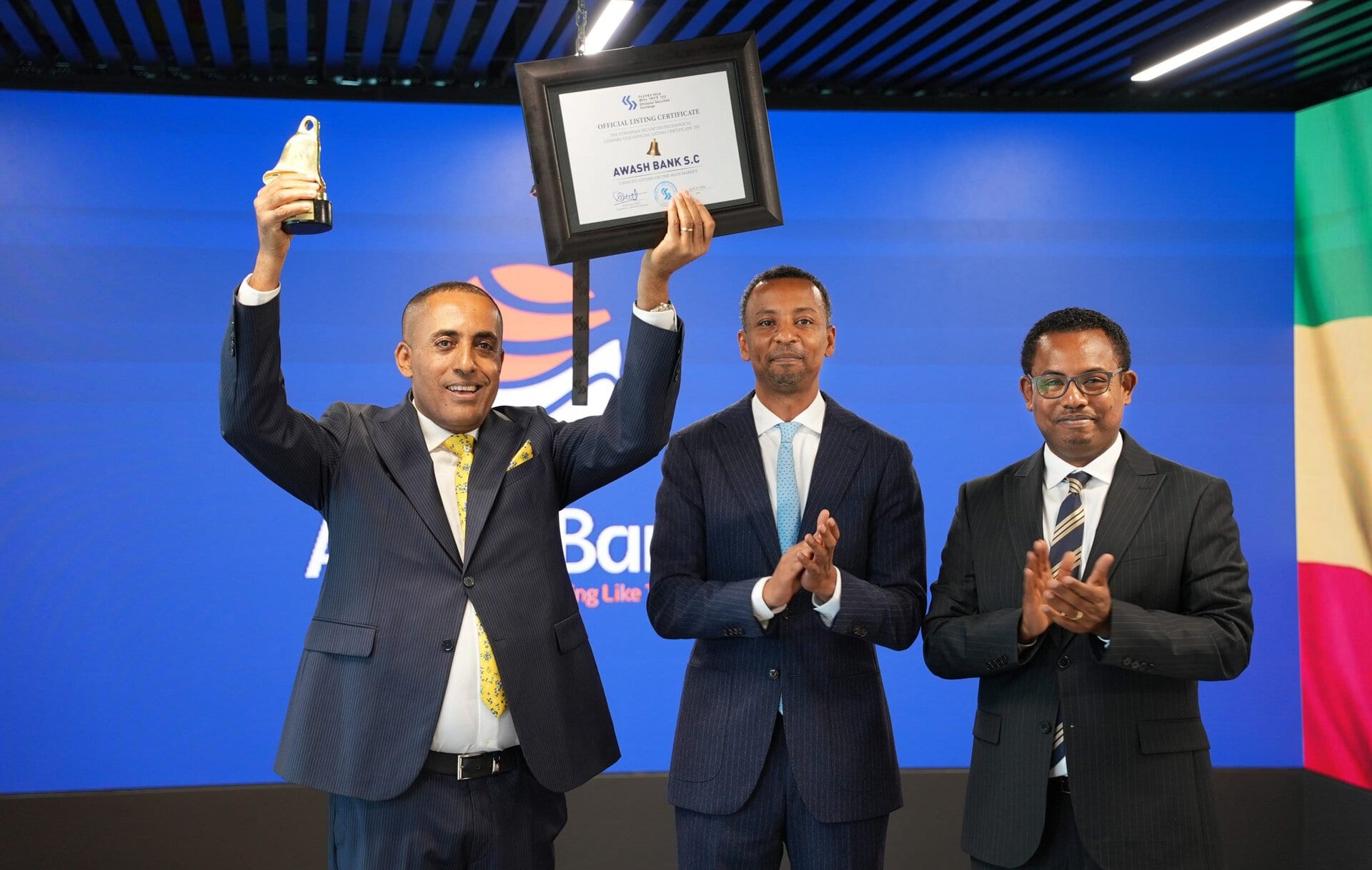 Three men on a stage holding up a certificate Celebrating the listing of Awash Bank SC on the ESX.