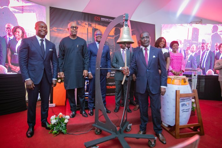 Men and women in formal wear around a ceremonial brass bell to celebrate the listing of Bridge Bank Cote d'Ivoire on the BRVM stock exchange.